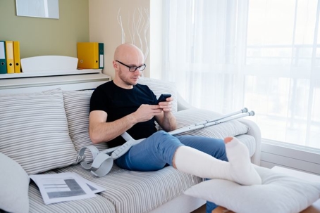 The man with the broken leg is sitting on the sofa at home.